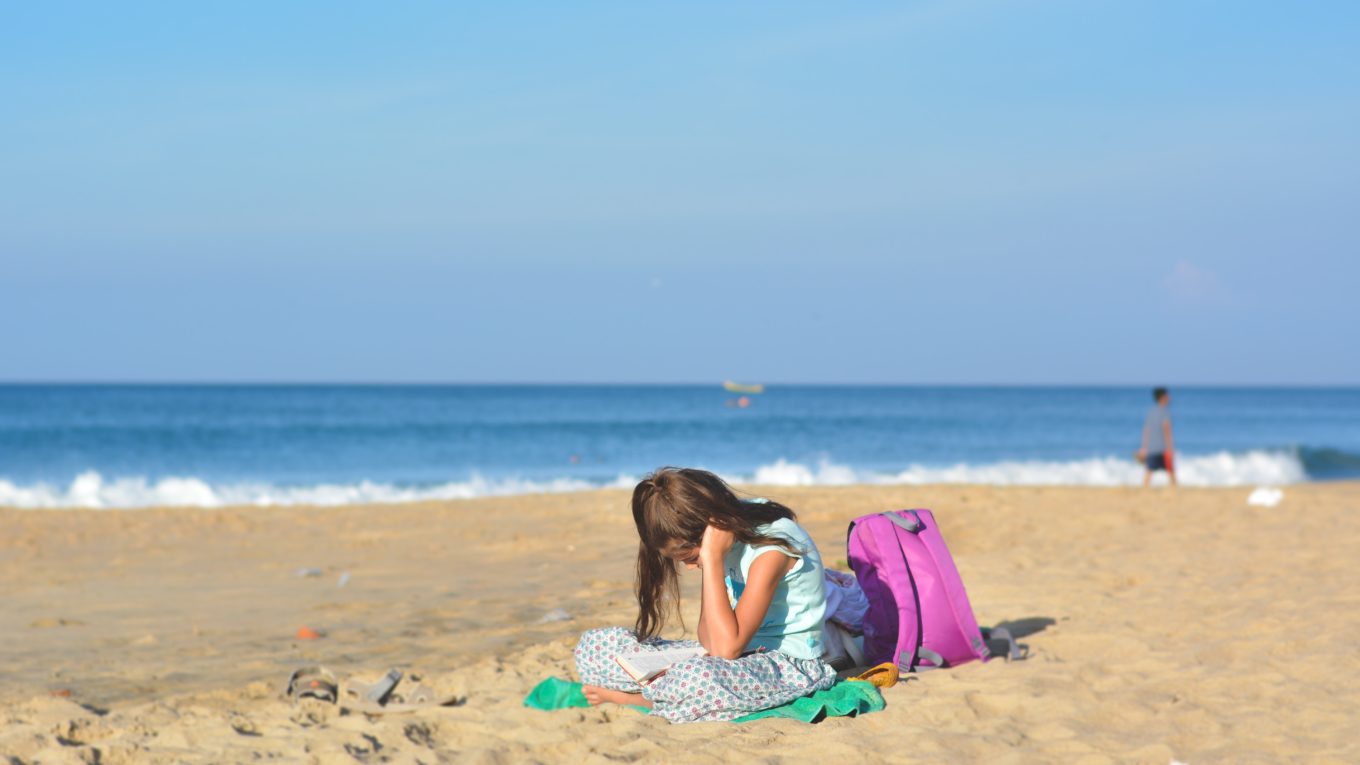 girl sits on shore near purple backpack under blue sky